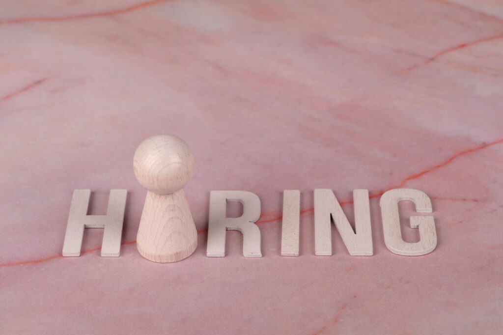 Wooden letters spelling 'hiring' on pink marble background with a peg.