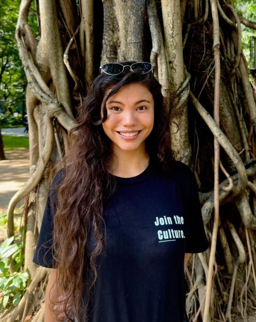Arielle Bustos, co-founder of TLC, smiling in a black Join the Culture t-shirt in front of a large fig tree